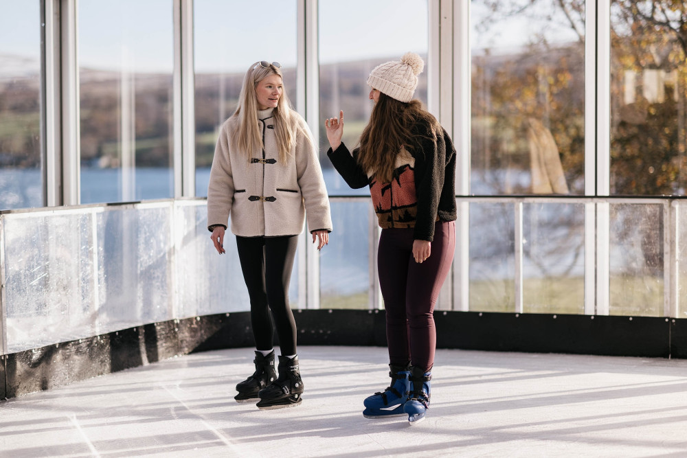 Two women ice skating with views over Ullswater lake