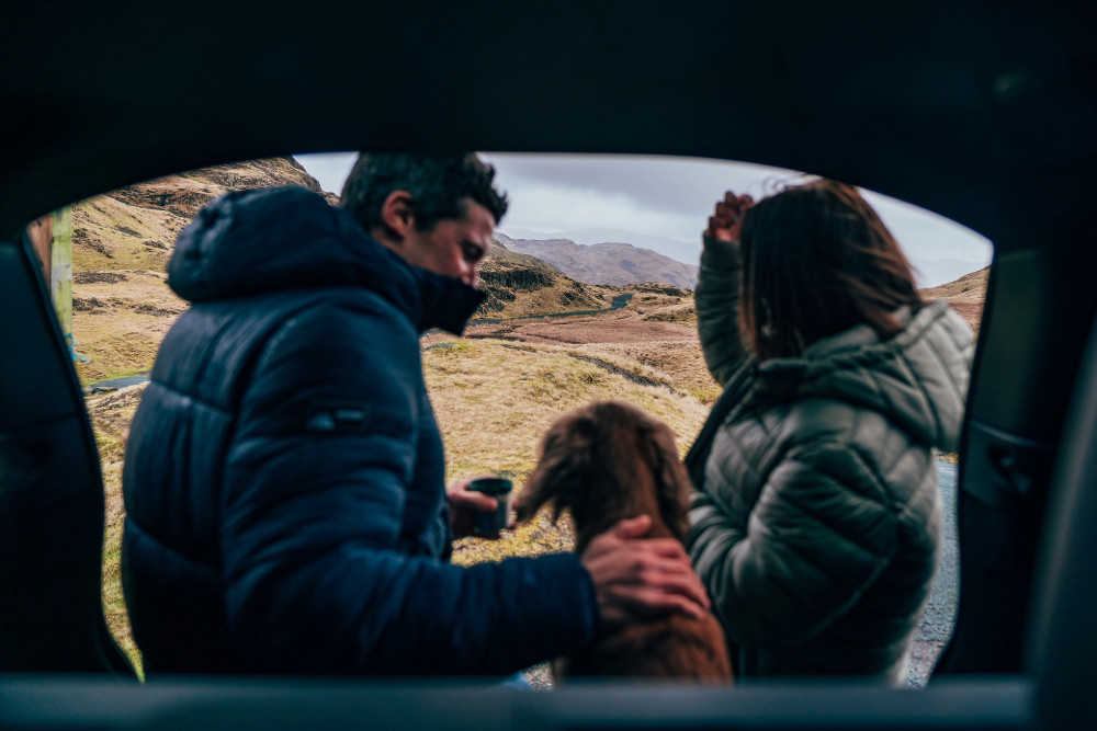 Two people sat in the boot of a land rover with their red cocker spaniel in the middle, looking out over the fells