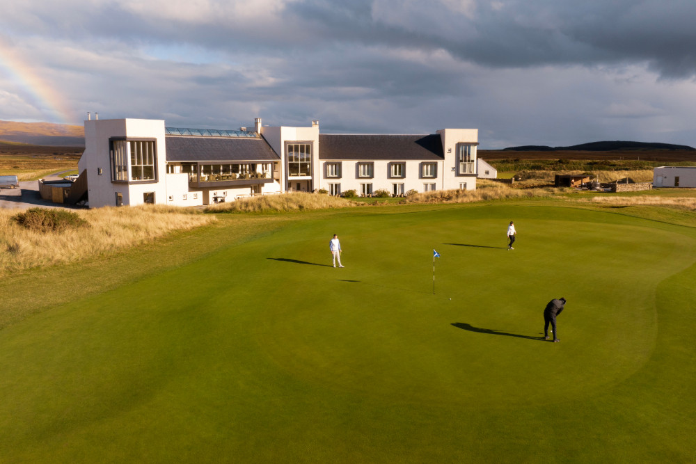 Aerial shot of three people playing the 18th hole of The Machrie, Islay golf course, with the hotel in the background and rainbow in the sky